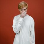 Young man in white sweater with hand on mouth standing against a red studio background.