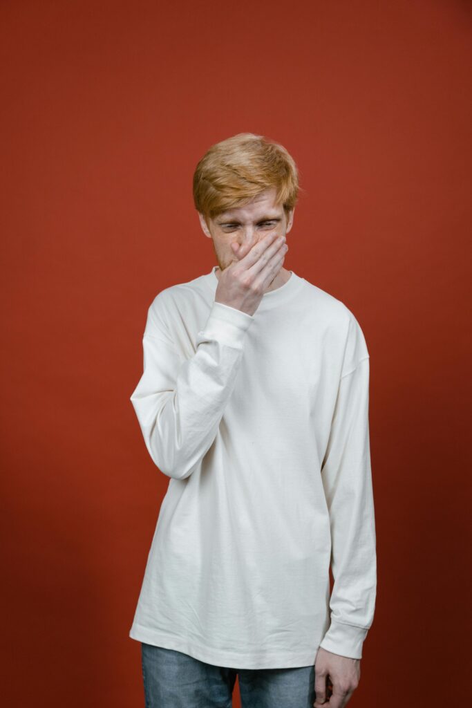 Young man in white sweater with hand on mouth standing against a red studio background.